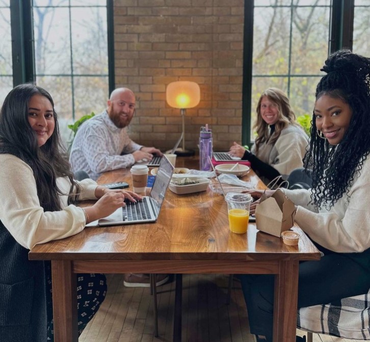 4 people sitting around a table, smiling at the camera. They have their laptops open in front of them and the table is covered with coffee, juices, and snacks.