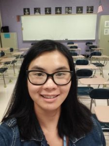 Samantha Gross, a Chinese woman in her early-20s, standing at the front of a high school classroom with student desks and a whiteboard visible in the background. Samantha has shoulder-length black hair and is wearing a blue jean jacket and black square glasses.