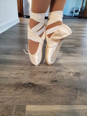 Close-up of Samantha Gross' feet standing en pointe. Samantha is wearing tan tights and pink satin pointe shoes with ribbons criss-crossing her feet and ankles.