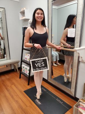 Samantha Gross, a Chinese-American woman in her mid-20s, standing en pointe in pink satin pointe shoes while holding onto a ballet bar with one hand. She is holding a sign in her other hand that says "I said yes to the shoes!" She has shoulder length black hair and is wearing a black leotard, black and white printed shorts, and pink tights.