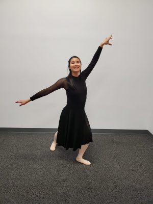 Samantha Gross, a Chinese-American woman in her mid-20s, posing gracefully in a lunge with her arms outstretched on a diagonal. She's wearing a long-sleeved black dance costume with a knee-length skirt and pink ballet shoes and tights.