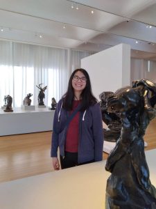 Samantha Gross, a Chinese-American woman in her mid-20s in a well-lit art gallery surrounded by dark bronze humanoid sculptures by Auguste Rodin. Samantha has long, black hair and black framed glasses. She is wearing a blue jacket over a red shirt.