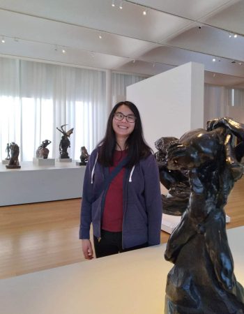 Samantha Gross, a Chinese-American woman in her mid-20s in a well-lit art gallery surrounded by dark bronze humanoid sculptures by Auguste Rodin. Samantha has long, black hair and black framed glasses. She is wearing a blue jacket over a red shirt.